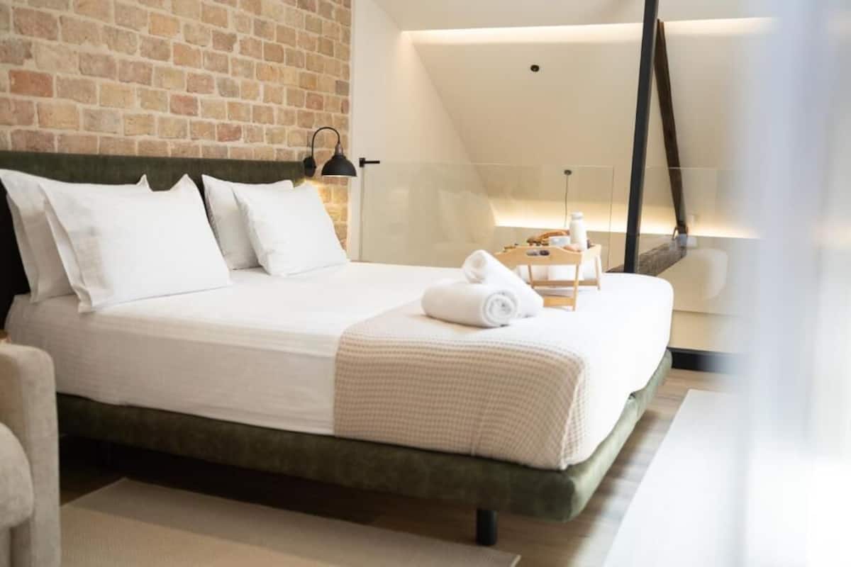 Une chambre d’hôtel moderne avec un mur en briques, un grand lit avec du linge de maison blanc, une table de chevet avec une lampe et une vue sur le ciel à travers une paroi en verre.