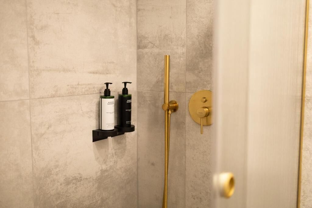 Un baño con ducha de oro, dispensador de jabón negro y una botella blanca sobre un estante negro.