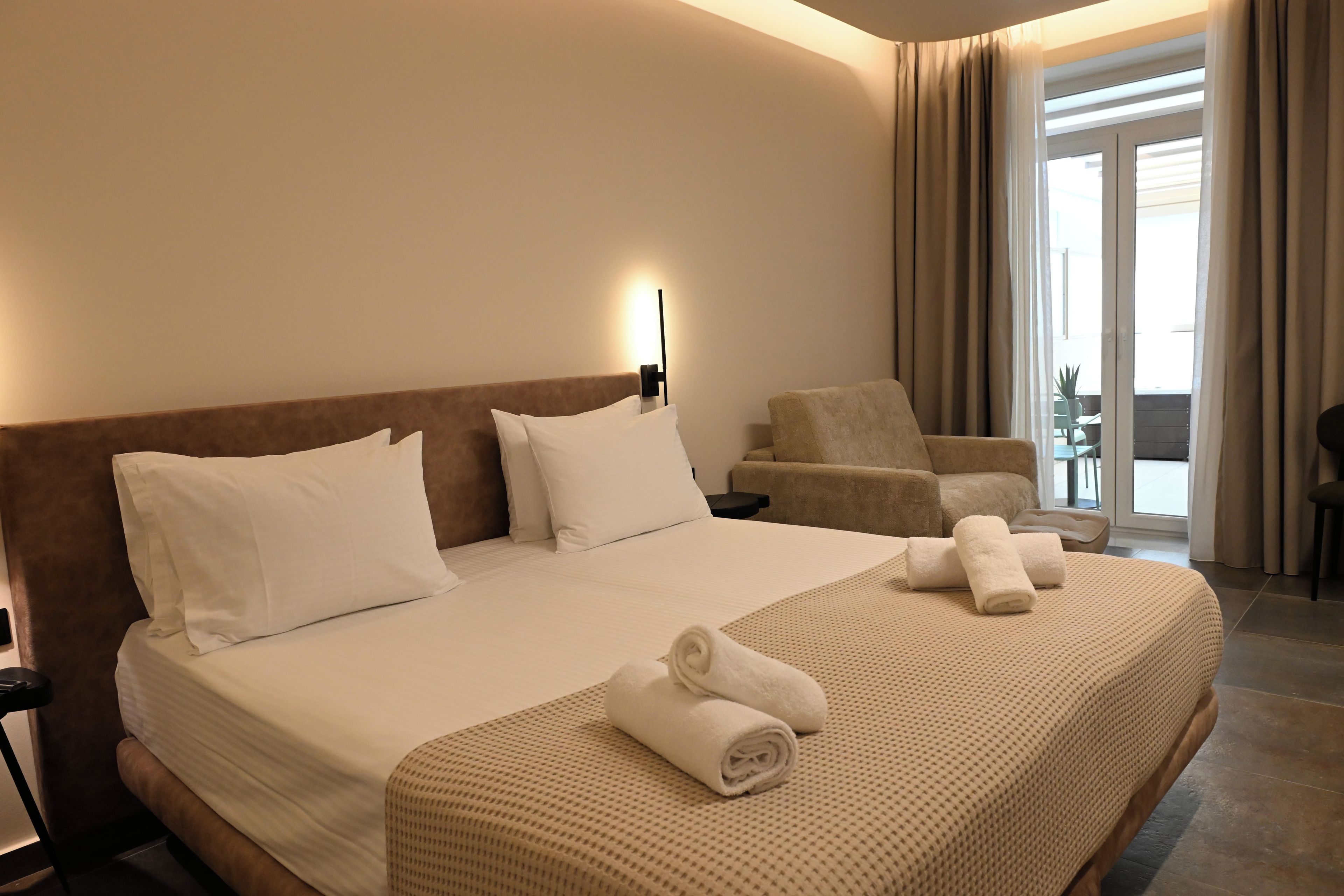 A hotel room with a large bed, two pillows, a nightstand, a chair, a table, and a window with curtains.