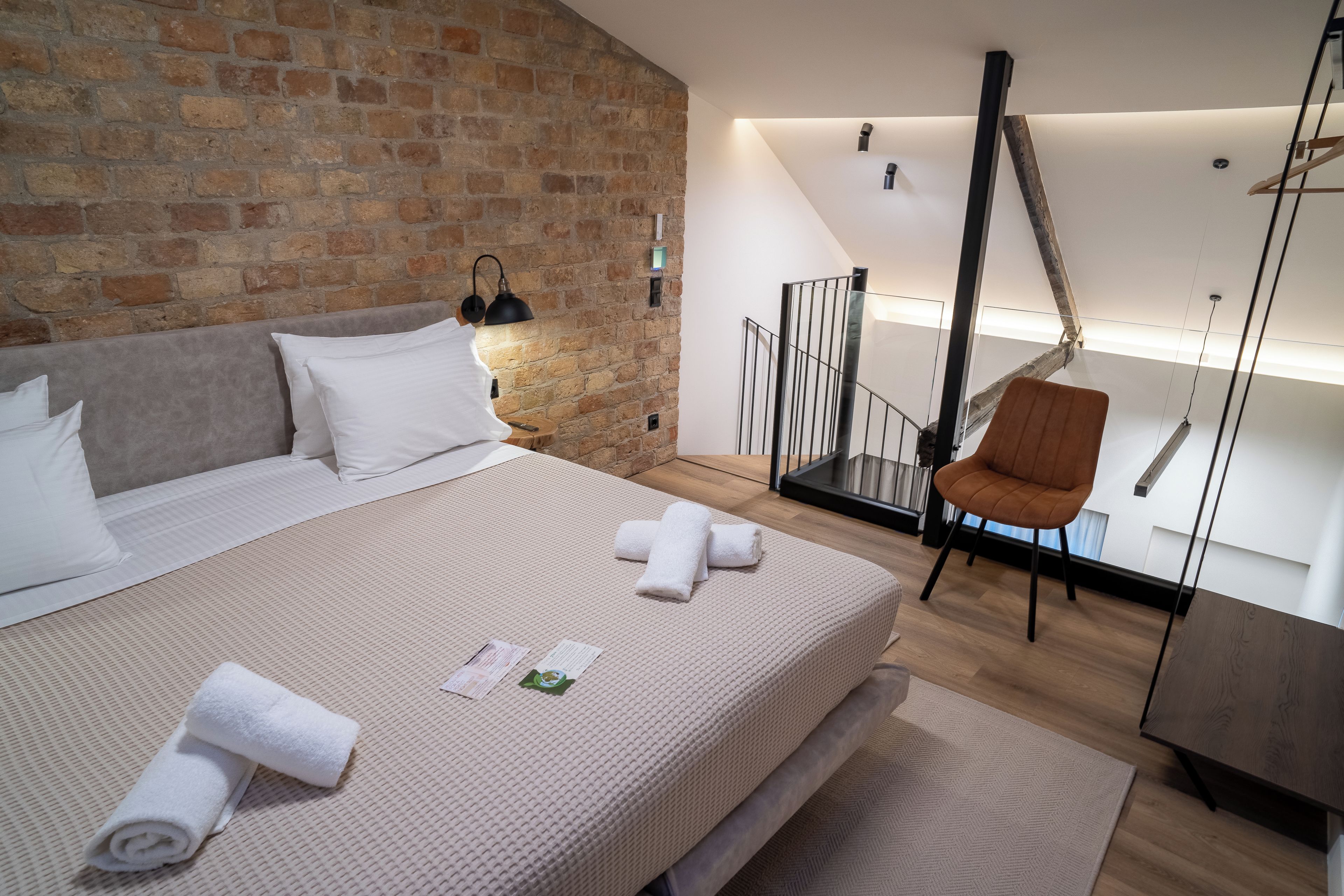 Una camera da letto moderna con parete in mattoni, un letto con lenzuola bianche, una sedia di legno e una scala che conduce a un livello superiore.
