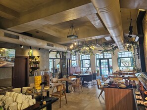 Interior de una cafetería con mesas, sillas, barra de café y ventanas amplias.