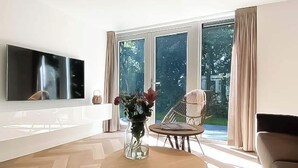 A modern living room with a flat-screen TV, a round coffee table with a vase of flowers, a wicker chair, and large windows with curtains. - Modern Coastal Retreat With Garden (Sint Maarten)
