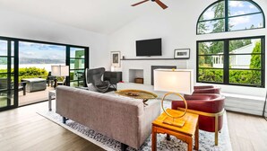 A modern living room with a sofa, armchairs, a TV, and large windows offering a view of greenery and a lake. - Greenbank Horizon Home | Water Views (Greenbank)