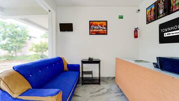 A reception area with a blue sofa, a small table, a fire extinguisher, and a wall-mounted television.