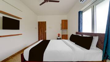 A hotel room with a bed, a ceiling fan, an air conditioning unit, a television mounted on the wall, and a window with curtains.