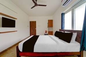 A hotel room with a bed, a ceiling fan, an air conditioning unit, a television mounted on the wall, and a window with curtains.