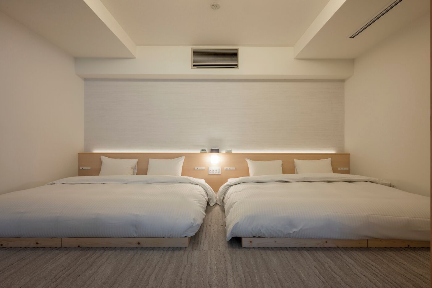 Une chambre double avec deux lits, une tête de lit en bois, une bouche d’aération au plafond et un luminaire au-dessus des lits.