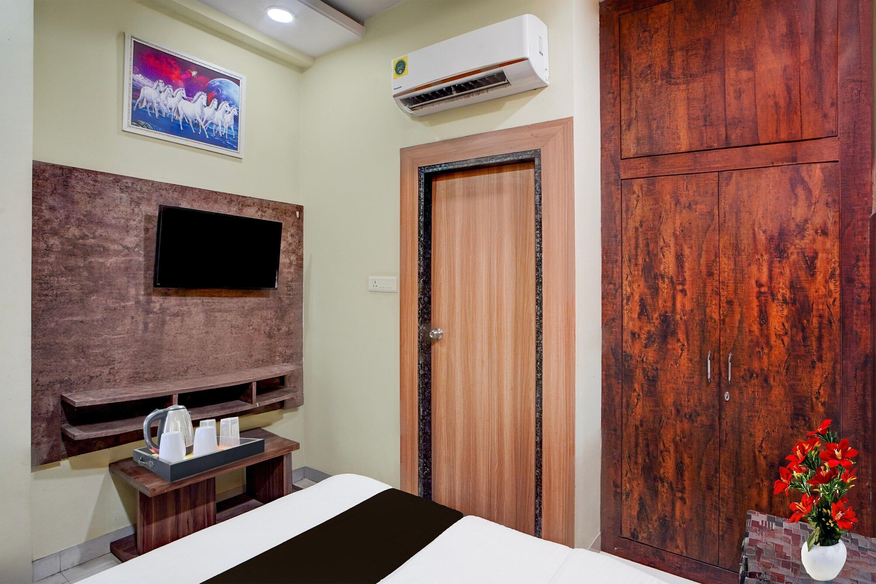 A bedroom with a bed, a TV mounted on a wall, a wooden wardrobe, and a small table with a kettle and cups.