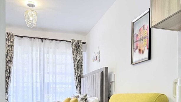 Et moderne soveværelse med seng, gul sofa, lille bord med plante, tekøkken og vindue med gardiner.