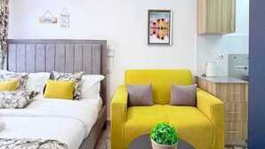 Et værelse med en seng, en sofa, et lille bord med en plante, et indrammet billede og et tekøkken.