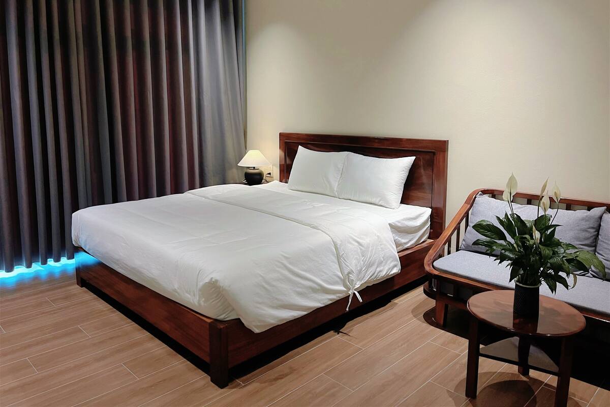 Habitación de hotel con una cama de madera, una mesita de noche con lámpara, una pequeña mesa redonda con una planta y vistas al exterior a través de una ventana con cortinas.