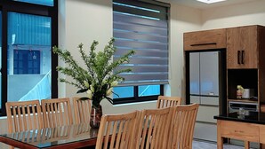 A dining area with a glass-top table, wooden chairs, a potted plant, a kitchen with a refrigerator, and a window with blinds. - An's Home Ninh Binh (Ninh Binh)