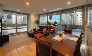 A modern living room with a sofa, dining table, and chairs, large windows with a view of buildings, and a small plant on the windowsill. - Terrace On Rose 2B2B 173m2 Pool Sauna/Steam Spa Room Gym FREE Secure Parking (Melbourne)