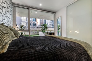 Une chambre moderne avec un grand lit, une vue sur la ville grâce à de grandes fenêtres du sol au plafond et un balcon agrémenté de plantes.