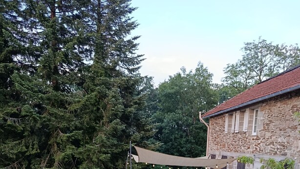 Un edificio de piedra con techo de tejas rojas, rodeado por un jardín de margaritas blancas y un área de descanso sombreada con sillas.