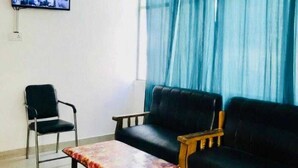 Una habitación con un sofá negro, un banco de madera, una silla de metal, una pequeña mesa, un ventilador de techo y un televisor montado en la pared.