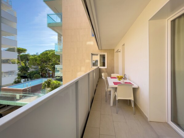 A balcony with a dining table, chairs, and a view of a swimming pool and greenery. - Modern flat in a prime location (Lignano Sabbiadoro)