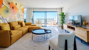 Ein modernes Wohnzimmer mit einem Ecksofa, einem runden Couchtisch, einem Fernseher und einem großen Fenster mit Aussicht.