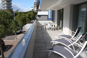 A balcony with white chairs and a table, overlooking a cityscape with modern buildings and trees. - Lignano stay with parking (Lignano Sabbiadoro)