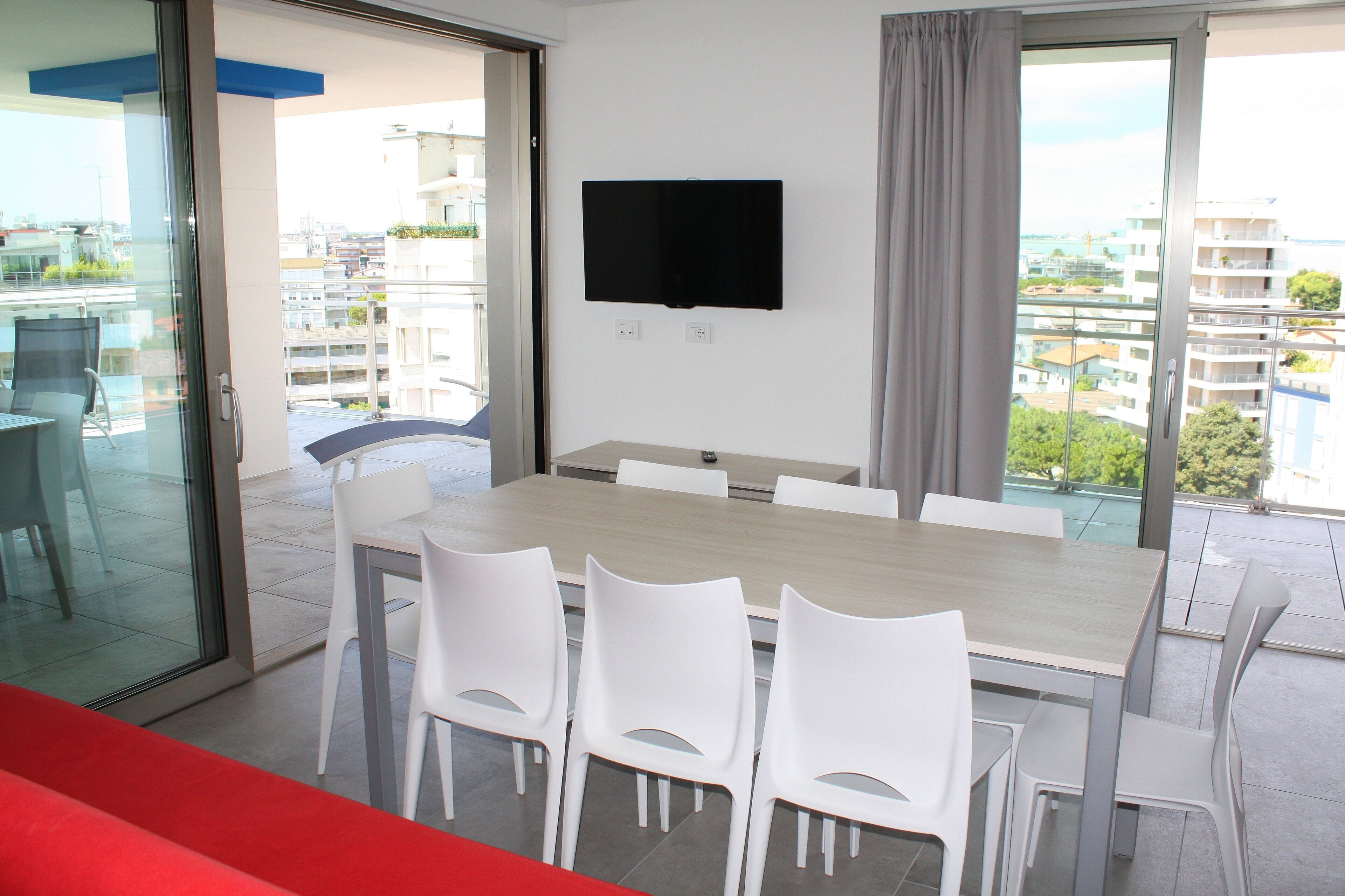 A modern dining area with a large table and white chairs, a flat-screen TV mounted on the wall, and a balcony with a view of the city.