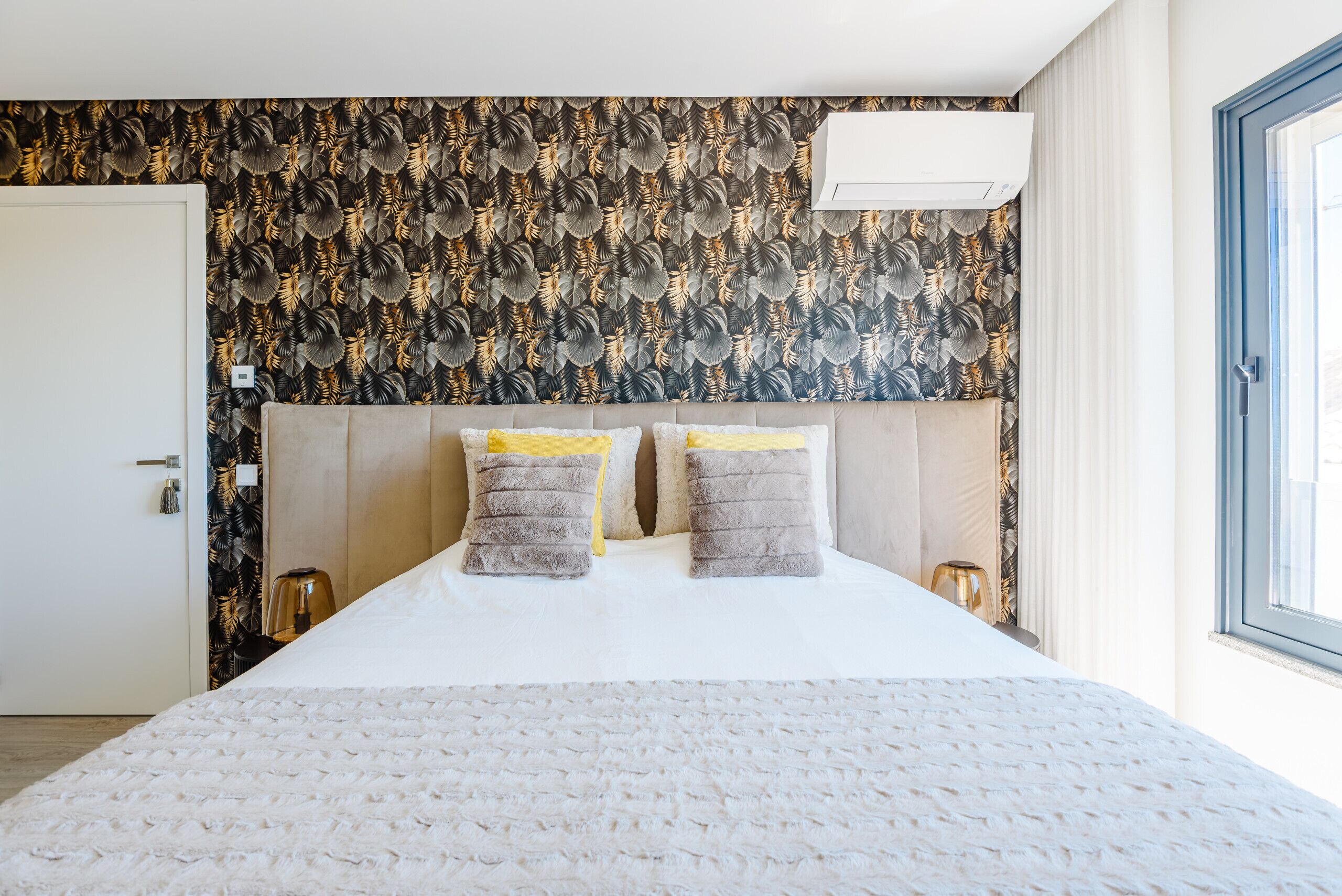 A hotel room with a large bed, a patterned wallpaper, a white air conditioning unit, and a window with curtains.