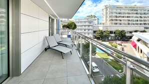 A balcony with two chairs, a glass railing, and a view of a cityscape with buildings and a car. - Bright apartment with balconies (Lignano Sabbiadoro)