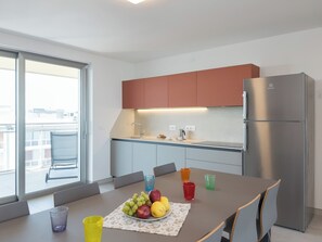 A modern kitchen with a built-in refrigerator, a dining table with chairs, and a view through a large window. - Lignano penthouse near beach (Lignano Sabbiadoro)