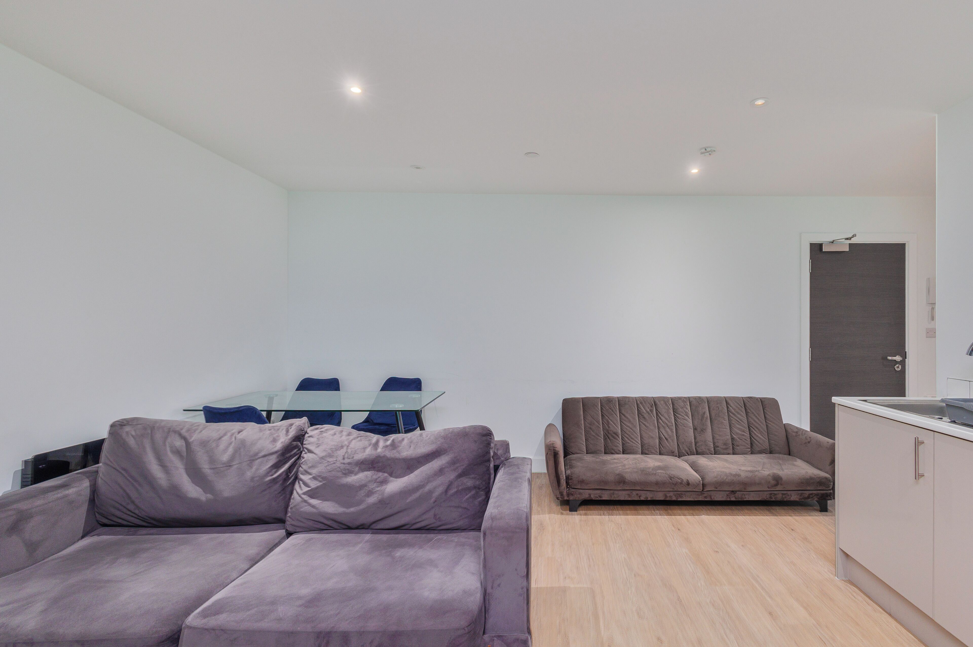 Een moderne woonkamer met een bank, een kleinere zithoek, een eettafel met glazen blad en stoelen, en een keuken met kasten.