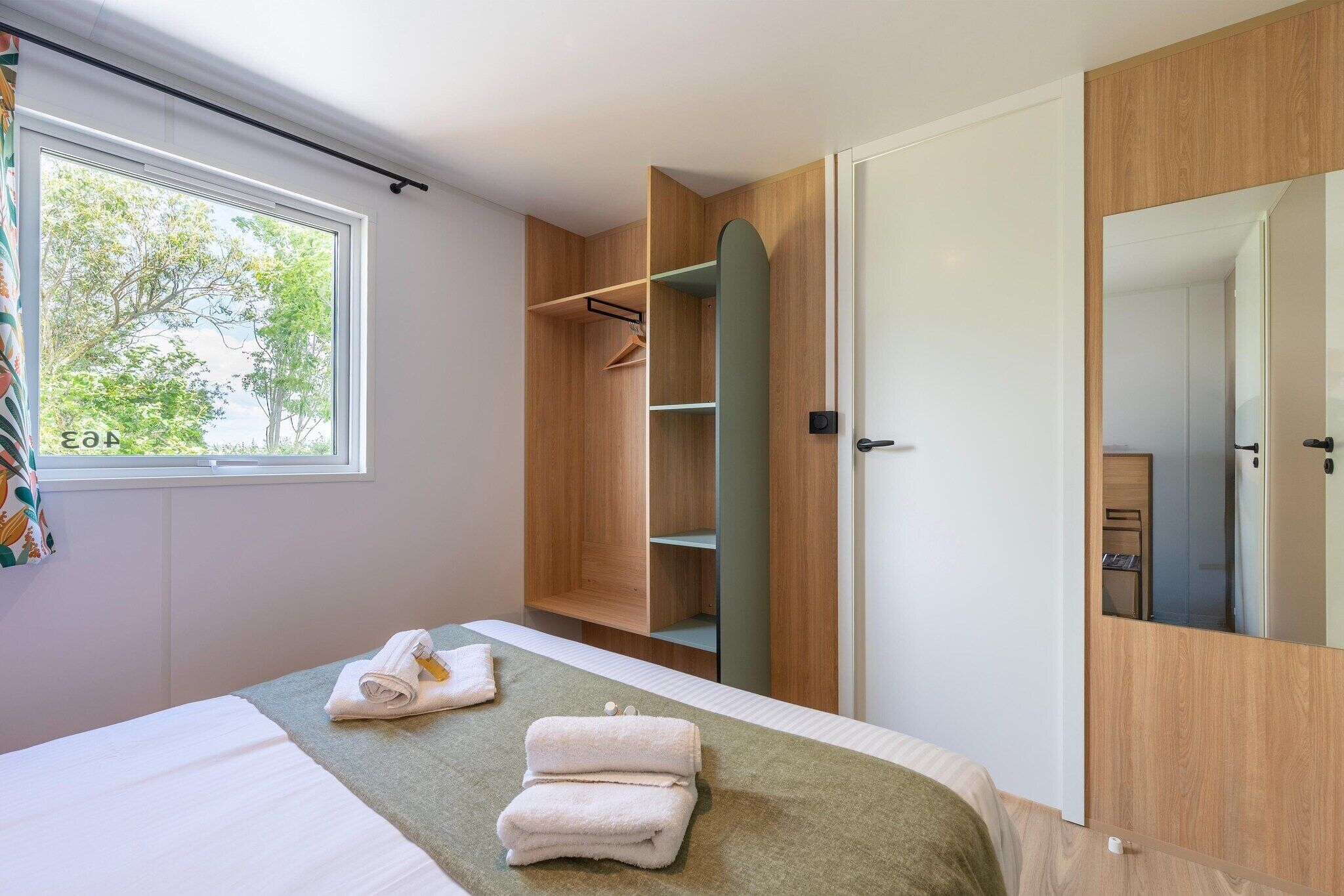 Une chambre d’hôtel avec un lit, une armoire, un miroir et une fenêtre donnant sur des arbres.