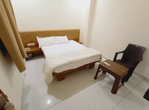A hotel room with a bed, a wooden nightstand, a chair, and a small table. - Hotel Shree Sanwariya Inn (Ujjain)