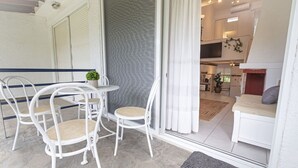 Una habitación con comedor que incluye sillas blancas y una mesita, una puerta corrediza de vidrio que da a una cocina, y una cama con cabecera roja.