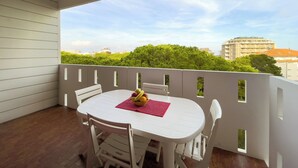 Un balcon avec une table et des chaises blanches, un panier de fruits et une vue sur des immeubles et des arbres.
