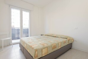 Quarto com cama, uma cadeira e porta de vidro deslizante que dá para uma varanda.