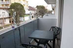 Varanda com mesa e cadeiras, vista para uma rua com edifícios e árvores.