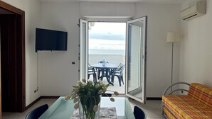 Een kamer met een eettafel, stoelen, een bank, een televisie en een open deur naar een balkon met uitzicht.
