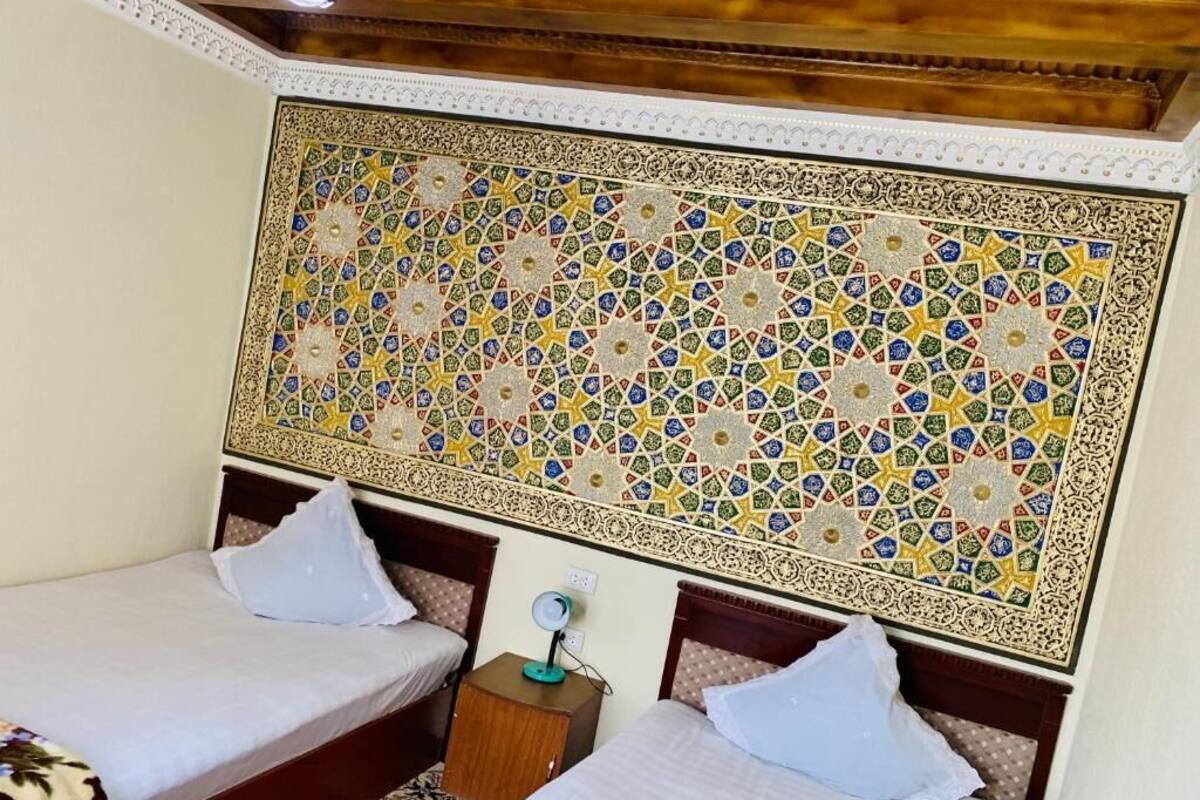 Habitación de hotel con dos camas, una mesita de noche con lámpara y un gran tapiz decorativo.