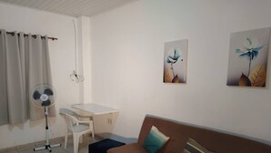 A room with a brown sofa, a small table, a chair, a standing fan, and two wall-mounted flower paintings. - 001 - Apartment Close to the Center (Vitoria da Conquista)