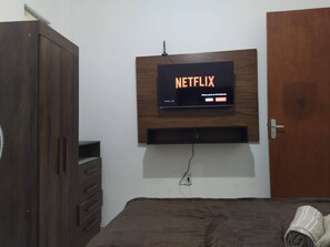 A room with a mounted television displaying the Netflix logo, a wooden wardrobe, a bed with a brown blanket, and a door.
