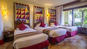 A hotel room with three beds, each with a red pillow and a patterned quilt. There are wall-mounted lamps and a window with a view of greenery. - Lake Manyara Serena Safari Lodge (Lake Manyara National Park)