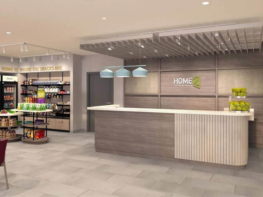 Uma área de recepção de hotel com design moderno, incluindo um balcão, prateleiras com lanches e uma placa que diz 'HOME 2 SUITES BY HILTON'.