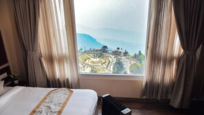 A hotel room with a bed, a radiator, and a window with a view of a town and mountains. - Anutri Hotels & Resorts  (Darjeeling)