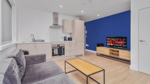 Una sala moderna con un sofá gris, una mesa de centro de madera, una zona de cocina con armarios blancos, una pared de acento azul y un televisor mostrando 'NETFLIX'.