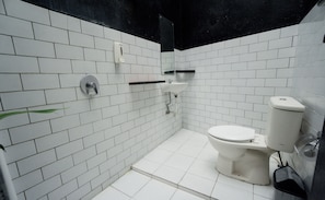 Une petite salle de bain aux murs recouverts de carreaux blancs, équipée de toilettes, d’un lavabo et d’un sèche-mains.