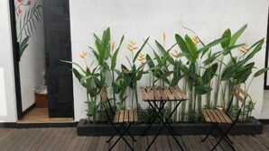 Una terraza de madera con una mesa y sillas de metal negro, rodeada de plantas verdes grandes y una pared blanca.
