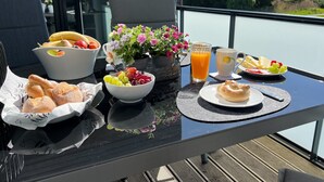 Una mesa con artículos para el desayuno, como frutas, pan y una bebida, con vistas a árboles y un edificio al fondo.