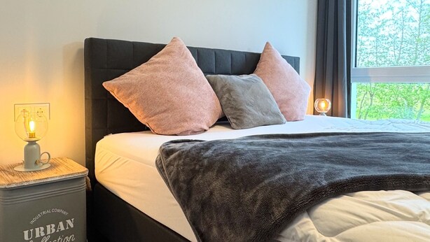 Un dormitorio con cama, almohadas, lámpara de noche y ventana con vistas.