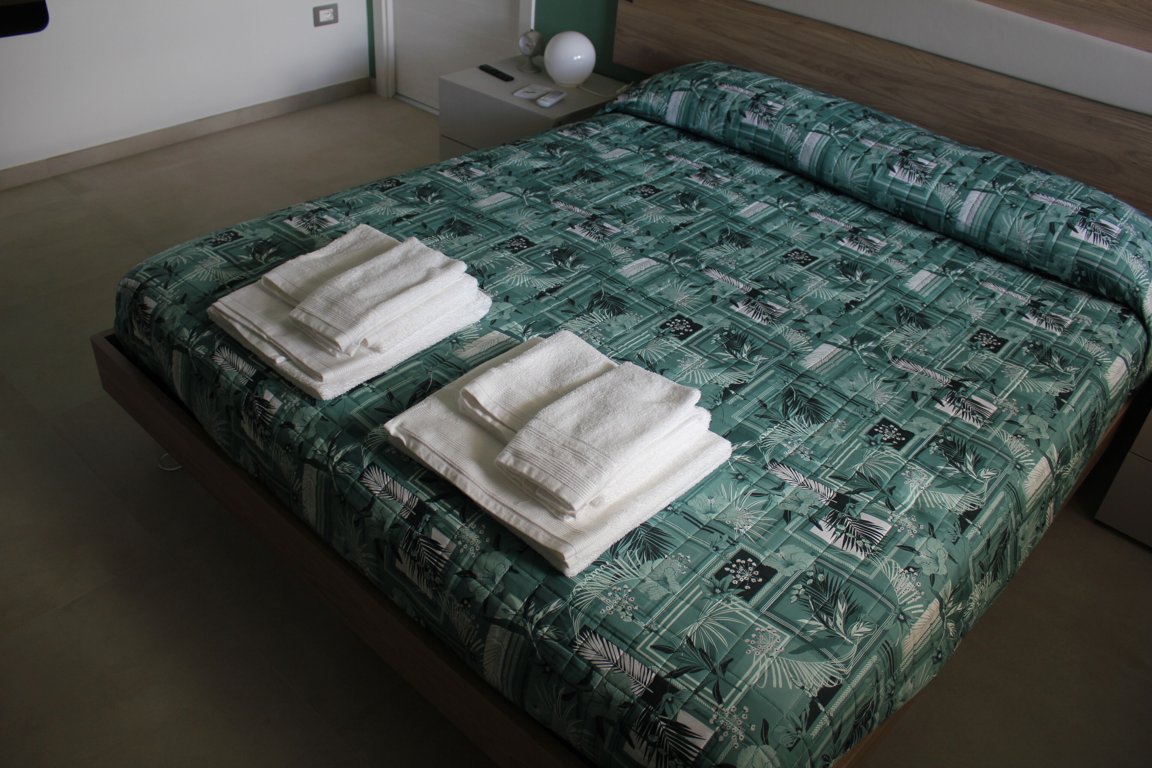 Un lit bien fait, recouvert d’une couvre-lit à motifs, deux serviettes blanches pliées et une table de chevet avec une lampe et un téléphone.