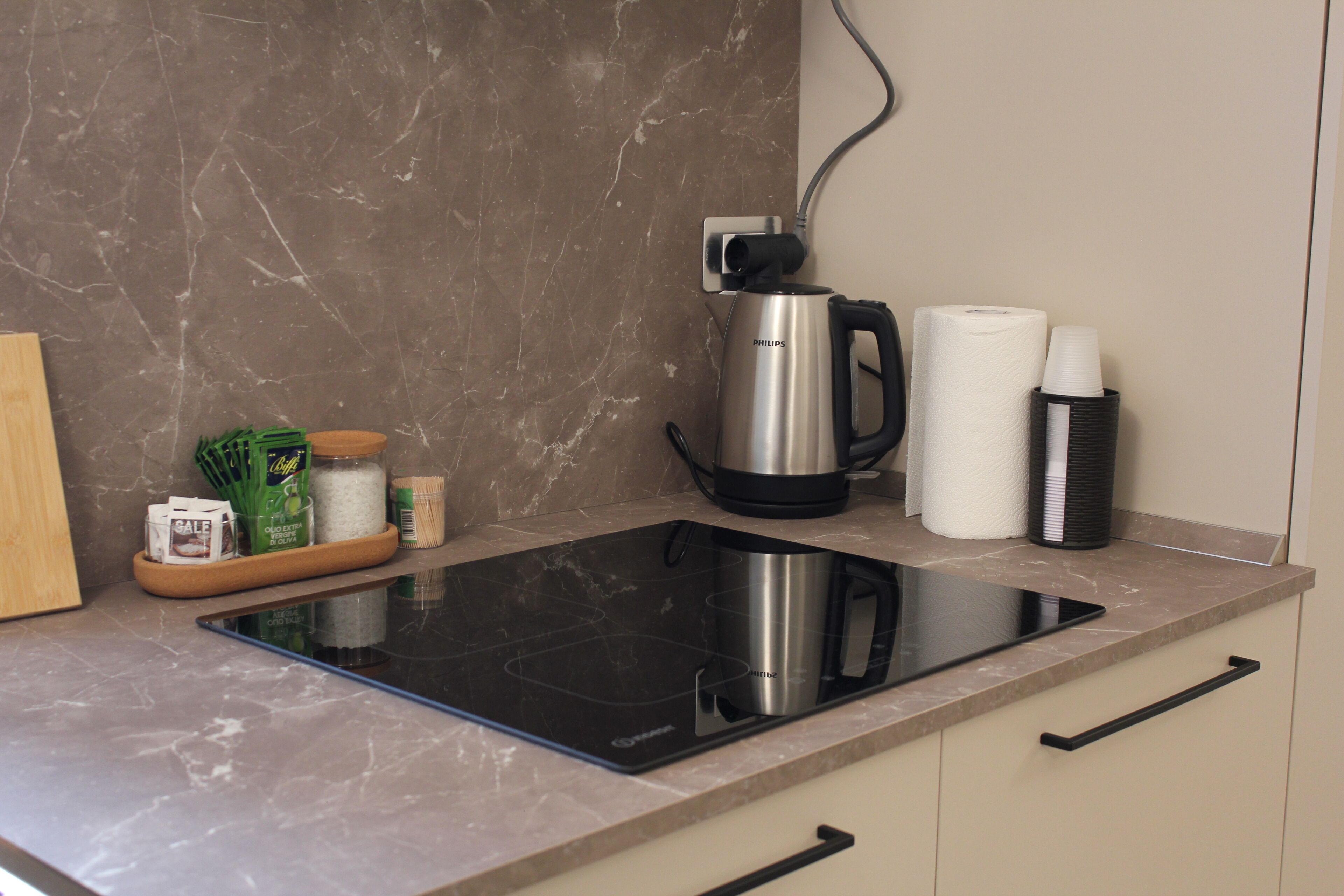 Una cocina con una placa de inducción negra, una cafetera de acero inoxidable, un rollo de toallas de papel y un termo negro.