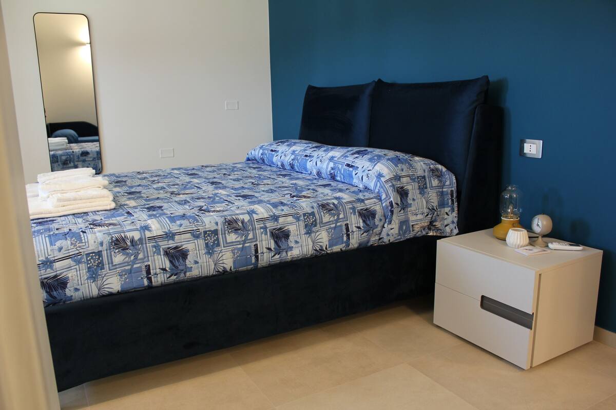 Un lit avec une couvre-lit à motifs, une table de chevet avec une lampe et un miroir au mur.