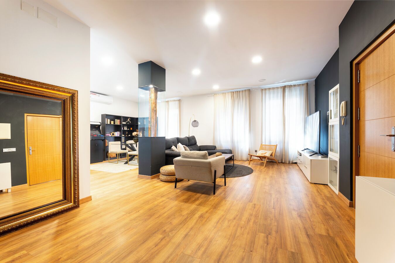 Una sala moderna con un sofá, sillones, una mesa de centro y un televisor. Se ve una estantería y un comedor al fondo.